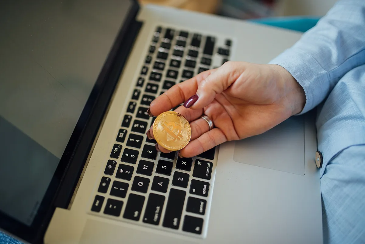 A physical bitcoin token held in a hand above a laptop.
