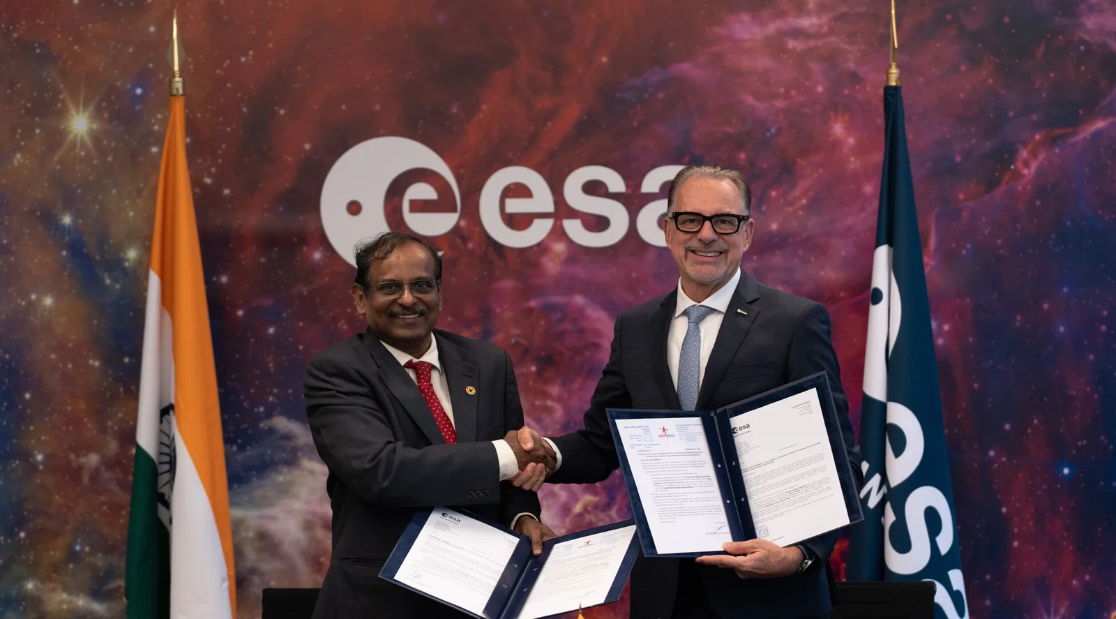 ISRO chairman V. Narayanan and ESA director general Josef Aschbacher shake hands in front of the Indian and ESA flags while holding signed documents.